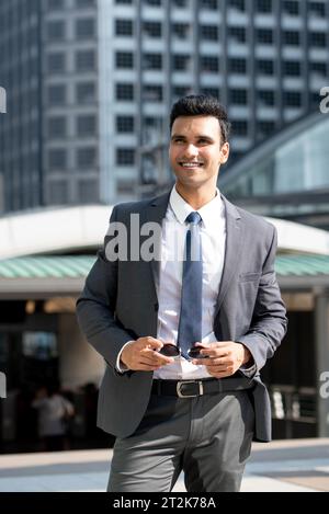 Ritratto di un elegante e sorridente uomo d'affari indiano che indossa una tuta grigia e regge gli occhiali da sole mentre si trova in piedi contro gli edifici della città Foto Stock