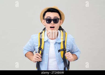 Scioccato giovane turista asiatico con zaino che indossa cappello e occhiali da sole che guardano la macchina fotografica, studio girato isolato su sfondo grigio Foto Stock