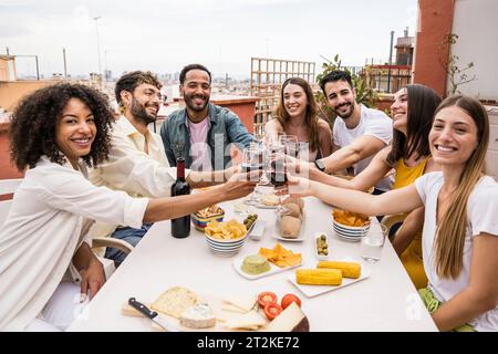 Un felice gruppo di amici che brindano con dei bicchieri da vino in terrazza guardando la macchina fotografica. Gruppo eterogeneo di giovani che bevono in una cena sul tetto Foto Stock