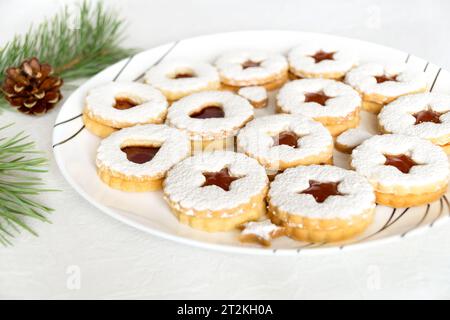 Biscotti al Linzer fatti in casa natalizi con marmellata cosparsa di zucchero a velo su piatto bianco. Atmosfera natalizia con rami di pino e cono di pino Foto Stock