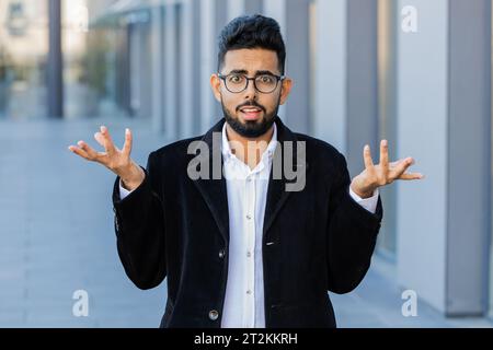 Cosa? Perché? Sorpreso un uomo d'affari indiano frustrato che alza la mano chiedendo il motivo del fallimento, dimostrando incredulità irritazione da problemi all'aperto. Confuso ragazzo indù arabo libero professionista in città Foto Stock