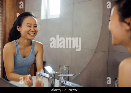 La giovane attraente donna asiatica con la parte superiore grigia si lava il viso e sorride al suo riflesso nello specchio rotondo. Affascinante ragazza coreana fa trattamenti all'acqua in m Foto Stock