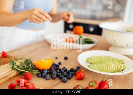 Una giovane ragazza sta preparando una colazione vegana in cucina. Primo piano di un piatto con avocado, rucola, frutti di bosco e agrumi su un tavolo di legno. TH Foto Stock