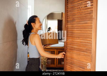 Bella giovane donna asiatica prende scatola di cartone fuori dall'armadio. Una donna coreana sorridente in abiti informali con pacchetto nel corridoio della casa. Attracti Foto Stock