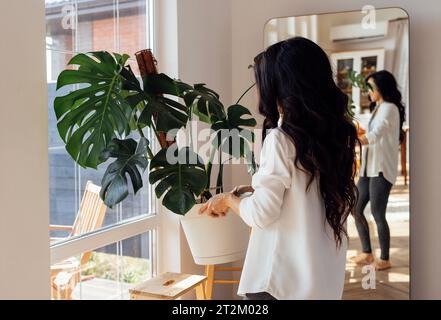 Un'attraente donna asiatica in abiti casual tiene una pentola con una pianta domestica. Un'affascinante giovane donna coreana si prende cura di un fiore monstera nei vivi Foto Stock