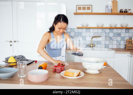 L'affascinante asiatica femminile prepara la colazione con frutti di bosco in cucina. Una giovane donna attraente fa dei panini. Una bella ragazza sorridente salvietta da tavolo a cena Foto Stock