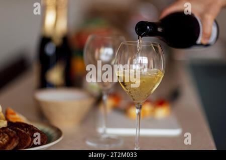 Primo piano di un elegante bicchiere con gambo lungo e champagne. Il processo di versamento del vino frizzante in un calice. Bottiglie di vino, piatti con spuntini Foto Stock