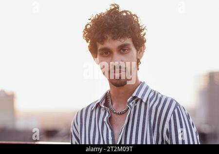 Primo piano di un ritratto maschile di un giovane di razza mista. Un attraente ragazzo dai capelli ricci con una camicia a righe sorride sullo sfondo di una grande città. Foto Stock