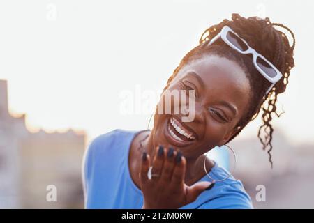 Una giovane donna africana ride e fa un bacio. Ritratto ravvicinato di un'affascinante donna sorridente dalla pelle scura con occhiali da sole. Una ragazza attraente e' l'havin Foto Stock