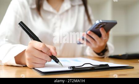 Donna asiatica che scrive e utilizza smartphone sul posto di lavoro. Foto Stock