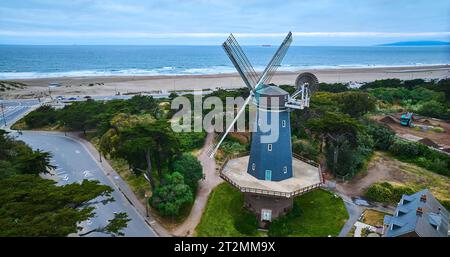 Mulino a vento Blue murphy circondato da alberi e strade che si affacciano sull'oceano blu con aerei da spiaggia sabbiosa Foto Stock