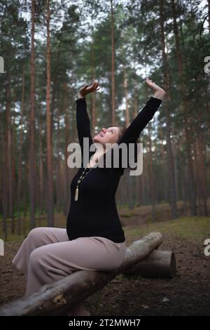 una donna incinta siede su un ceppo nella foresta e alza le mani. Foto Stock