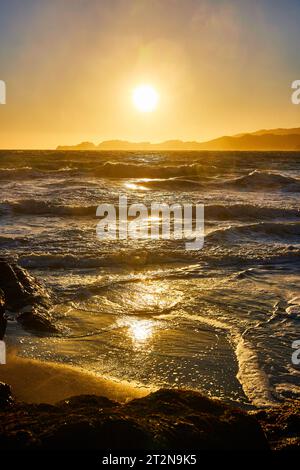 Glorioso tramonto dorato sulle onde dell'oceano che si snodano sulla spiaggia sabbiosa con montagne lontane Foto Stock
