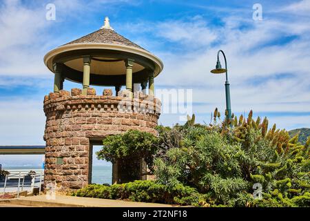 Vista sull'oceano attraverso la finestra della torre in pietra marrone con piante accanto al marciapiede sotto il cielo blu nuvoloso Foto Stock