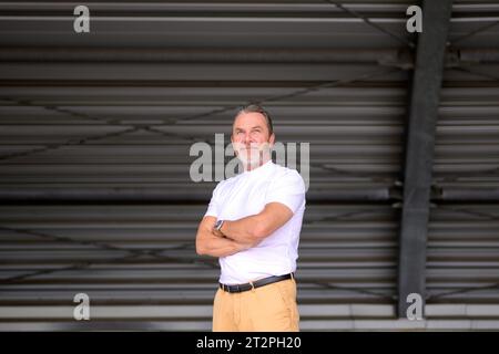 Ritratto del corpo a tre quarti di un attraente uomo dai capelli grigi con una T-shirt bianca con le braccia incrociate, indossa un orologio elegante e sembra amichevole, Foto Stock