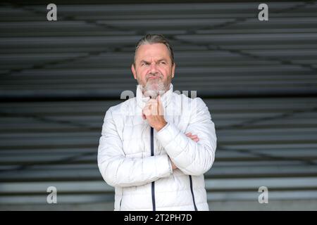 Attraente uomo dai capelli grigi che indossa una giacca invernale bianca con una mano sulla giacca che sembra scontroso lateralmente, davanti a una parete in ferro ondulato Foto Stock