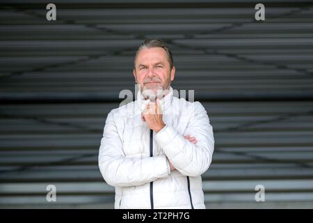 Attraente uomo dai capelli grigi che indossa una giacca invernale bianca con una mano sulla giacca che sembra amichevole con la fotocamera, davanti a una parete in ferro ondulato Foto Stock