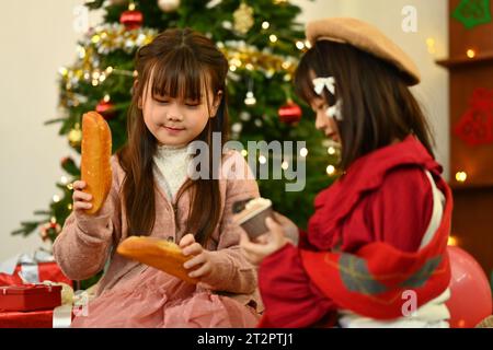 Adorabili sorelline che mangiano pane fresco e muffin in soggiorno la vigilia di Natale Foto Stock