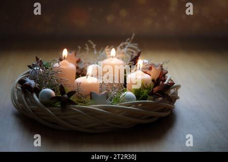 Corona dell'avvento in tonalità naturali realizzata in vimini con candele beige, muschio e decorazioni natalizie su un tavolo di legno su sfondo scuro, copia spac Foto Stock