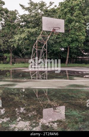 Il tabellone da basket in legno con il cerchio in metallo e la rete bianca mostrano il loro riflesso sull'acqua piovana su tutto il vecchio cemento da basket Foto Stock