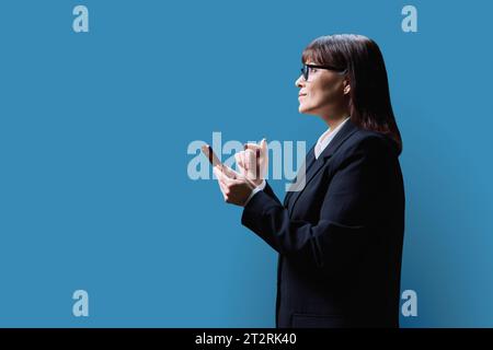 Donna d'affari matura con smartphone, vista del profilo su sfondo blu Foto Stock