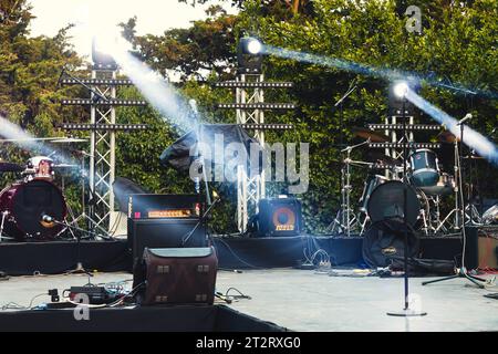 Un palco da concerto vuoto con attrezzature musicali, amplificatori per chitarra, illuminati da faretti mobili, in un parco pubblico di giorno Foto Stock