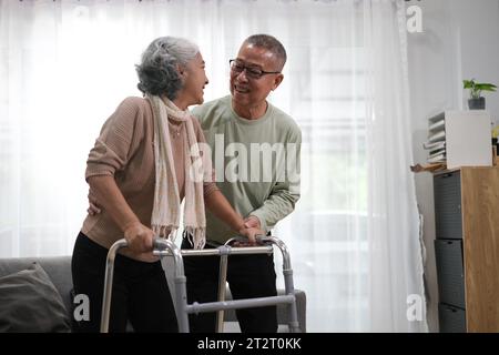 Un uomo anziano che aiuta sua moglie con un telaio che cammina al chiuso. Foto Stock