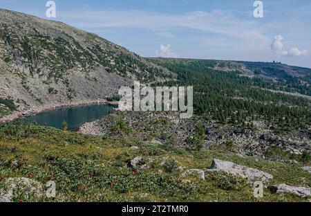 Colline cosparse di grandi pietre, un lago blu sottostante, alberi di conifere nella foresta verde e rocce all'orizzonte. Paesaggio estivo di natura selvaggia a bassa quota Foto Stock