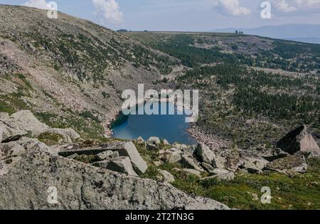 Un lago di montagna verde e blu circondato da colline di pietra con ripidi pendii, rocce, enormi pietre e foresta di conifere. Vista dall'alto del paesaggio faunistico estivo Foto Stock