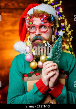 Buon Natale. Buon anno nuovo. Uomo barbuto con cappello di Babbo Natale e occhiali da festa con piccolo albero di Natale. Uomo serio con barba decorata e capelli tinti Foto Stock