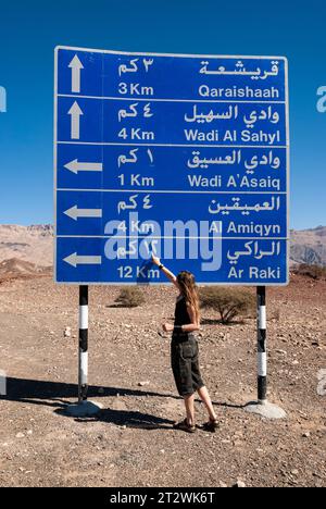 Una donna indica un cartello stradale scritto in arabo sulle montagne dell'Oman Foto Stock