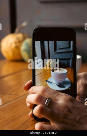 Donna che fa una foto al caffè mattutino. Tazza di caffè con zucche sullo sfondo sullo schermo del telefono. Telefono cellulare in mano donna. Concetto di blog. Foto Stock