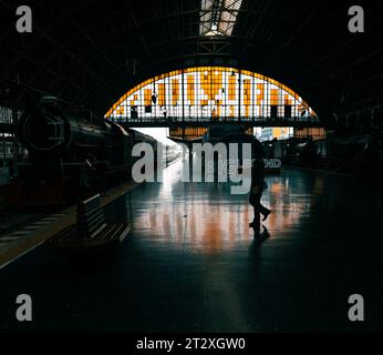 Uomo che cammina su un binario della stazione ferroviaria verso un vecchio treno a vapore. C'è un posto a sedere in primo piano e una finestra di vetro colorato sullo sfondo. Foto Stock