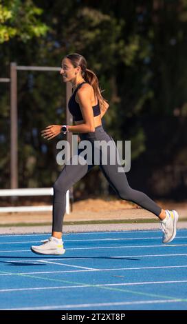 Bellissima giovane runner con abbronzatura sottile, vestita con abbigliamento sportivo aderente, con un'espressione felice e una corsa sorridente con stile e una tecnica perfetta Foto Stock