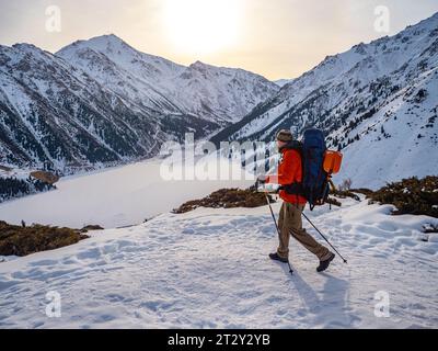 Un viaggiatore uomo fa trekking in montagna in inverno. Un uomo con un piumino rosso e uno zaino cammina sullo sfondo di un mou congelato Foto Stock