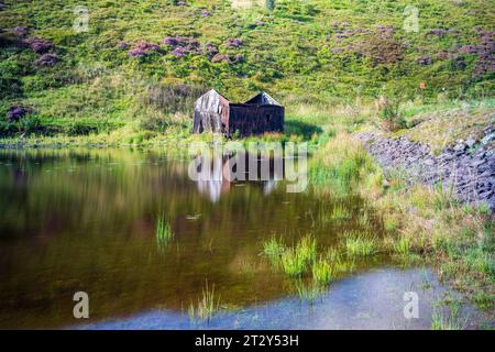 Una rovina di una baracca ai margini di un lago in Scozia in una soleggiata mattinata estiva durante una mattinata estiva Foto Stock
