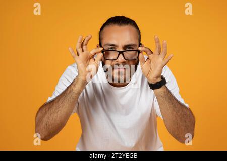 Uomo dalla barba millenaria europea con una t-shirt bianca che regola meticolosamente gli occhiali con i bordi neri Foto Stock