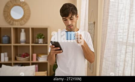 Bellissimo giovane ispanico si gusta tranquillamente il caffè mattutino a casa, completamente immerso nei messaggi sul suo smartphone, comodamente seduto nella sua vita Foto Stock