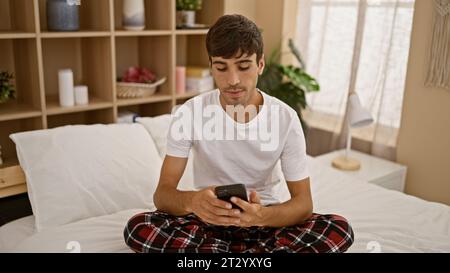 Giovane ispanico, vestito in pigiama, si rilassa sul suo comodo letto, immerso nel scrivere un messaggio serio sul suo smartphone nell'accogliente atmosfera di hi-Fi Foto Stock