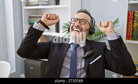 Uomo di mezza età sicuro di sé con i capelli grigi, un lavoratore professionale sorridente che indossa le cuffie, che celebra la sua vittoria in ufficio: Un vincitore nella sua i professionale Foto Stock