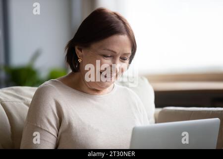 Donna anziana degli anni '60 che usa un computer portatile seduto sul divano a casa Foto Stock