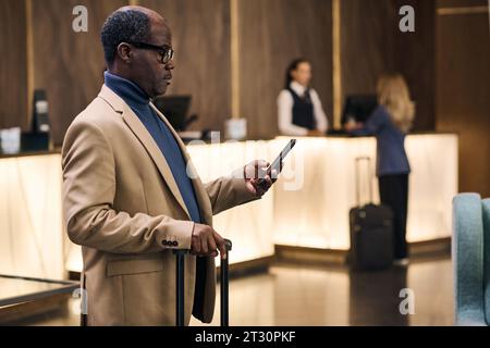 Uomo d'affari afroamericano che controlla la sua sistemazione online sullo smartphone mentre si trova in piedi con i bagagli in hotel Foto Stock