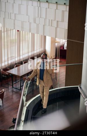 Vista ad alto angolo di un uomo d'affari afroamericano che parla al telefono mentre si è in piedi sulle scale nella hall dell'hotel Foto Stock