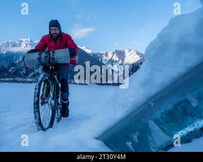 In inverno, un uomo va in bicicletta in montagna. Gita in bicicletta invernale. Pattinaggio sul ghiaccio di un lago di montagna ghiacciato. Mattina presto d'inverno in alto Foto Stock