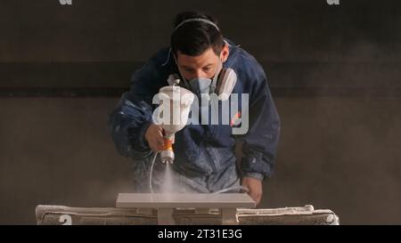 Il lavoratore dipinge pannelli di legno con pistola a spruzzo. Creativo. Il lavoratore dipinge prodotti in legno con pistola a spruzzo in officina. Officina di verniciatura per la pittura del legno Foto Stock