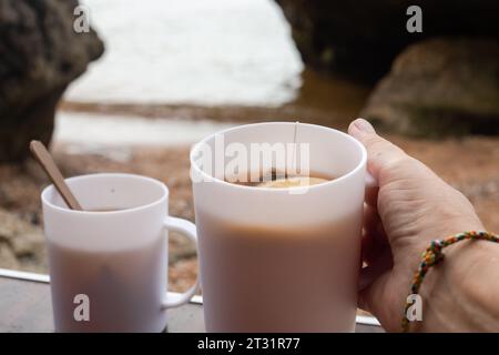 La mano di un uomo tiene una tazza bianca di tè su una costa rocciosa in autunno. Viaggi e turismo. Picnic nella natura. Foto Stock