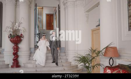 L'uomo e la sposa scendono le scale. Azione. Gli sposi novelli in abiti scendono dall'ufficio del registro. Sposi e sposi scendono le scale dell'ufficio del registro Foto Stock