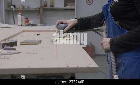 La donna macina il prodotto in legno in officina. Creativo. Donna in grembiule lucida il telaio in legno. Officina di falegnameria con legname da donna Foto Stock