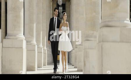 Adorabili sposi che passeggiano lungo le colonne del parco. Azione. Uomo in completo e donna in abito bianco corto vicino a colonne di marmo. Foto Stock