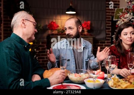 Persone diverse che si godono le vacanze della vigilia di natale a casa, sedute intorno a un tavolo festivo e mangiano cibo delizioso. La famiglia e gli amici si divertono a condividere ricordi, festeggiare l'evento di Natale. Foto Stock